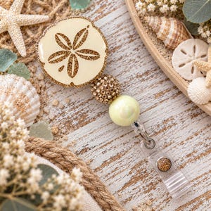 May include: A badge reel featuring a cream-colored sand dollar disc with a gold glitter floral pattern. The design includes a gold rhinestone bead, a smooth pale yellow bead, and a clear plastic strap with a gold glitter accent button.