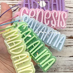 May include: A set of five custom name tags made of glittery resin with scalloped edges. Each tag features raised lettering in a solid color, including the names Abigail, Genesis, Savanna, Catalina, and Madaline, each attached to a thin colored cord.