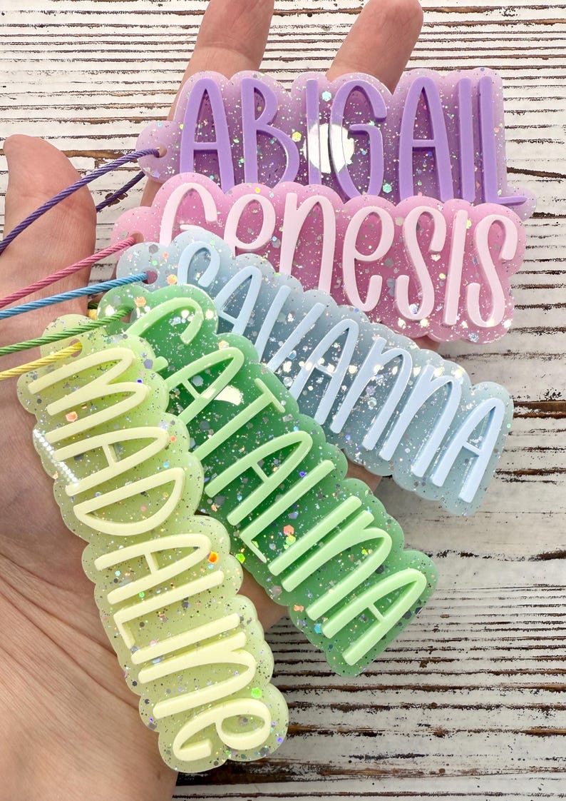 May include: A set of five custom name tags made of glittery resin with scalloped edges. Each tag features raised lettering in a solid color, including the names Abigail, Genesis, Savanna, Catalina, and Madaline, each attached to a thin colored cord.