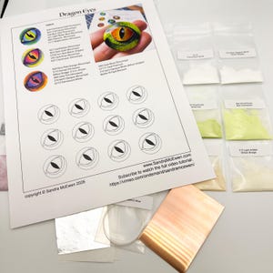 May include: A craft kit with a printed guide titled "Dragon Eyes" featuring eye designs and color swatches. Small bags of powder pigments and metal sheets are also present, along with wire and other supplies.