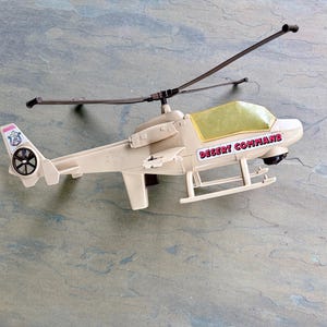 May include: Tan toy helicopter with the words "Desert Command" printed on the side. The helicopter has a yellow tinted windshield and a black rotor. The tail features a circular emblem with a black propeller.