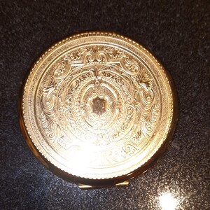 Op de afbeelding: Een vergulde compact spiegel met een sierlijk, gegraveerd ontwerp. De spiegel heeft een scharnierend deksel en een ronde vorm.