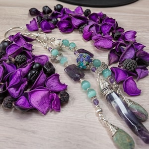 May include: A handmade keychain with silver-toned metal, featuring a variety of beads in shades of teal, purple, and white. The keychain has two long, purple stone pendants with green accents. The background includes purple flower petals and black berries.