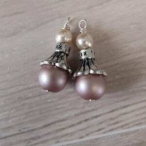 May include: A pair of dangle earrings featuring a light pink, pearlescent bead at the base, topped with a silver-colored decorative element and a smaller, cream-colored bead. The earrings have a simple hook for pierced ears.