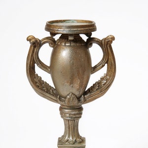May include: A gold-toned metal candle holder with an ornate design. The holder has a round top and two handles. The base is square with intricate details.