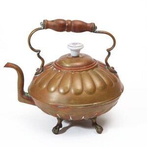 May include: A vintage copper tea kettle with a wooden handle and a porcelain knob. The kettle has a fluted design and sits on three feet.