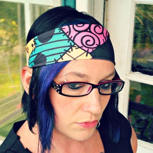 May include: A woman with blue hair is wearing a black and white headband with a colourful patchwork design. The headband has a pink, yellow, green, and blue pattern. The woman is also wearing glasses.