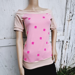 Puede incluir: Top rosa claro con hombros descubiertos y mangas cortas. El top presenta un escote y mangas en rosa más claro, y un panel central con un patrón de cuadrados rosa brillante. La parte inferior del top es de color rosa claro.