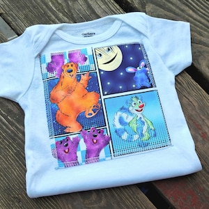 May include: White baby bodysuit with a colourful graphic featuring cartoon characters. The design includes a bear, a rabbit, and other creatures in a comic book style. The bodysuit is made of soft material and has short sleeves.