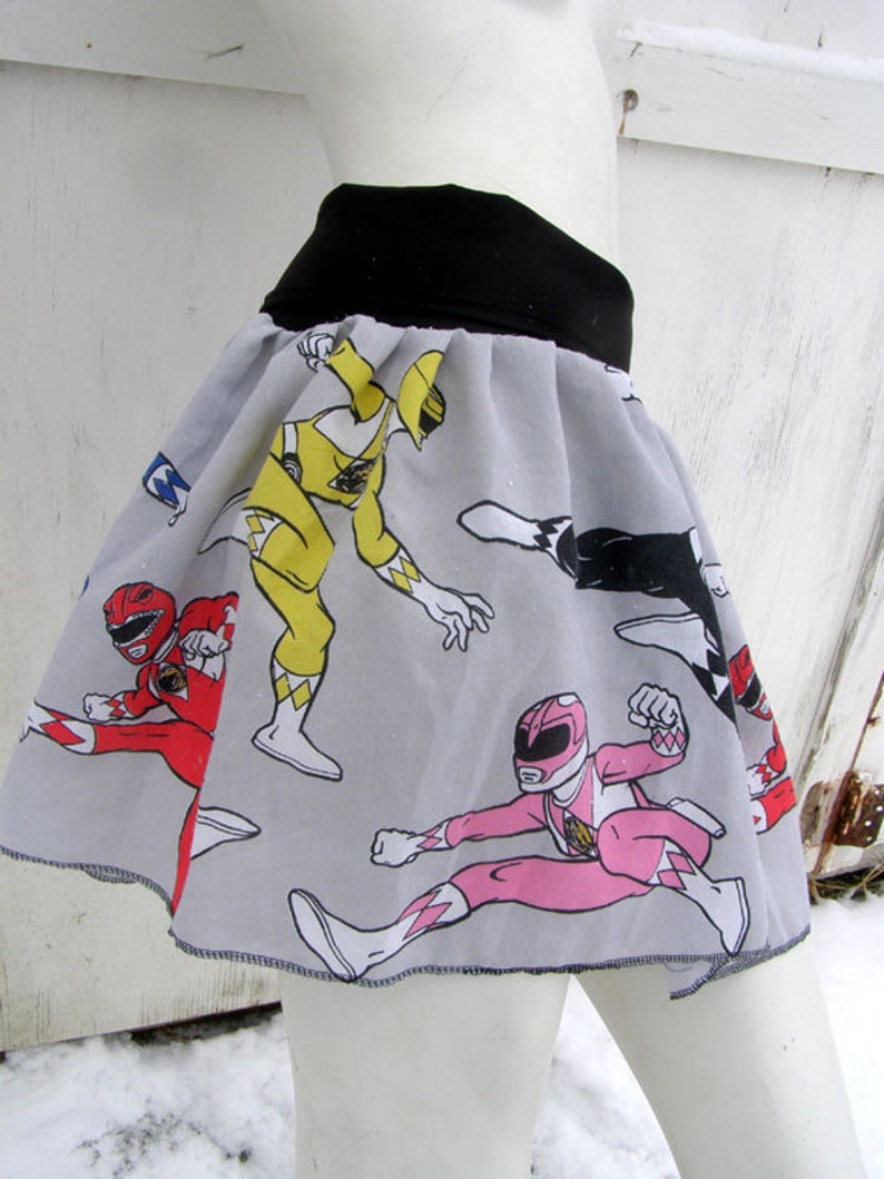 K&ouml;nnte beinhalten: Ein grauer Rock mit einem schwarzen Bund und einem Aufdruck der Power Rangers. Der Rock zeigt die gelben, roten, pinken und schwarzen Power Rangers.