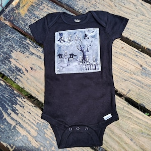 May include: A black short-sleeved baby bodysuit featuring a square patch on the chest. The patch displays a monochromatic graphic illustration of skeletons, black cats, and a bare tree in a graveyard setting. The garment has a round neckline and snaps.