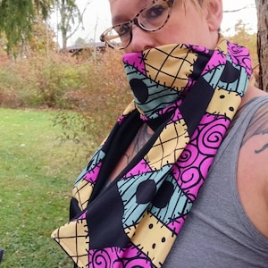 May include: A black infinity scarf with a colorful patchwork design featuring a stitched pattern, swirls, and black dots. The scarf is made of a soft, stretchy fabric and is perfect for adding a touch of whimsy to any outfit.
