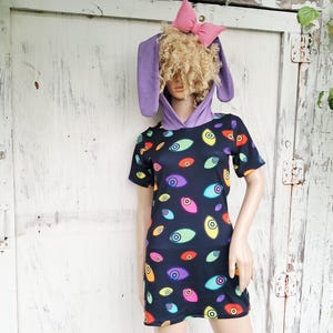 May include: A short-sleeved dress featuring a black background with a repeating pattern of colorful eyes in shades of red, yellow, purple, and teal. The dress includes an attached hood with long purple rabbit ears and a pink bow on top.