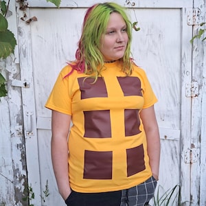 May include: A bright yellow short-sleeved t-shirt featuring a graphic design of six dark brown squares arranged in two vertical columns of three, creating a grid-like pattern across the front of the garment.
