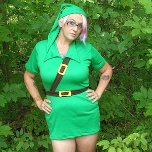 May include: A woman wearing a green tunic with a brown belt and a brown strap across her chest. She is wearing a green hat with a pointed tip. She has pink and purple hair and is standing in a woodland setting.