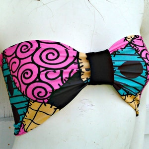 May include: A vibrant bandeau top with a patchwork design, showcasing pink swirls, teal stripes, and yellow diamond patterns. It features black fabric and a central black band. This distinctive top is a playful addition to any wardrobe.