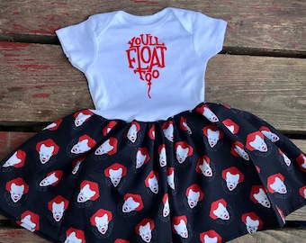 Puede incluir: Un body blanco para bebé con texto rojo que dice "You'll Float Too" en forma de globo. El body se combina con una falda negra con un patrón repetido de una cara de payaso.