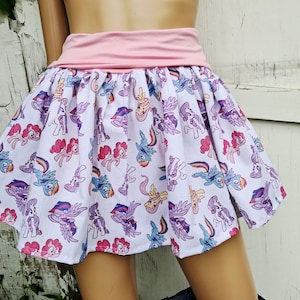 Op de afbeelding: Een witte rok met een roze tailleband en een patroon van kleurrijke tekenfilmpony's. De pony's zijn in verschillende poses en kleuren afgebeeld, waaronder roze, paars, blauw en geel. De rok is gerimpeld en heeft een uitlopende vorm.