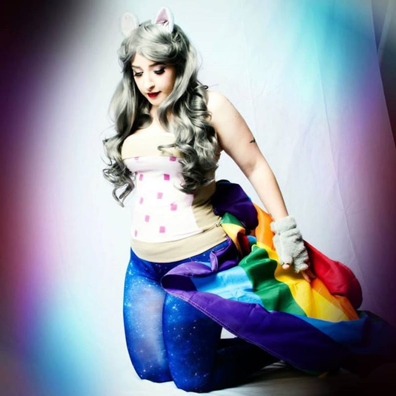 May include: A woman wearing a white and pink corset with a rainbow flag draped around her waist. She is wearing blue leggings with a galaxy print and has grey hair with cat ears.