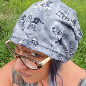 May include: A gray beanie with a black and white skeleton pattern. The beanie has a white zig-zag stitch around the edge. The beanie is being worn by a person with glasses.