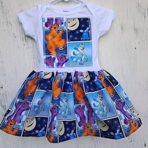 May include: A white baby dress featuring a cartoon graphic on the bodice and skirt. The graphic depicts a bear, a rabbit, and other characters in a comic book style. The skirt is blue with a repeating pattern of the characters.