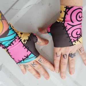 May include: A pair of black, pink, yellow and blue fingerless gloves with a patchwork design. The gloves have a stitched pattern and are inspired by the character Sally from the film The Nightmare Before Christmas.