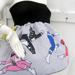 K&ouml;nnte beinhalten: Ein graues und wei&szlig;es tr&auml;gerloses Kleid mit einem schwarzen Oberteil und einem Aufdruck der Power Rangers. Der Aufdruck zeigt die Red, Blue, Black und Pink Rangers.