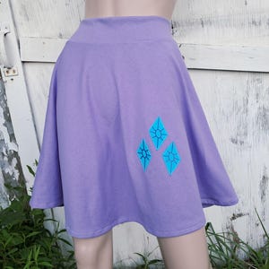 May include: A lavender skater skirt with a wide waistband. The skirt features three turquoise diamond-shaped appliques. The skirt is made of a soft fabric and is suitable for casual wear.