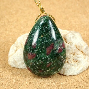 Puede incluir: Un colgante de piedra preciosa verde en forma de lágrima con inclusiones de rubí rojo. El colgante está enhebrado en una cadena de oro.