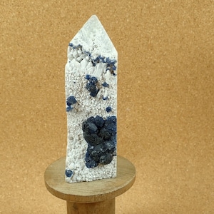 Op de afbeelding: Een witte kwartskristalpunt met blauwe en zwarte insluitsels. Het kristal staat op een houten standaard.