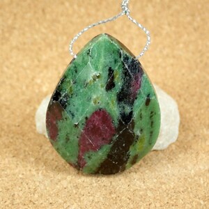 May include: A teardrop-shaped pendant made of green, black, and pink zoisite. The pendant has a silver cord attached to it.