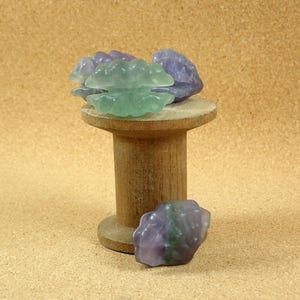 Puede incluir: Tres cristales de fluorita verde y púrpura sobre un carrete de madera. Los cristales están agrupados y tienen una textura natural y rugosa.