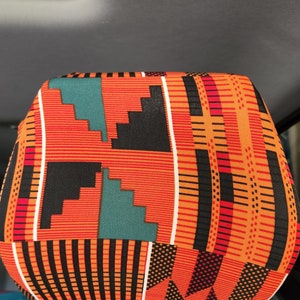 May include: A colorful headrest cover with a geometric pattern in shades of orange, black, white, red, and teal. The pattern is reminiscent of African Kente cloth.