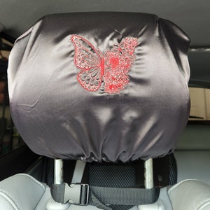 May include: Black satin car headrest cover with a red embroidered butterfly design. The cover has a black strap with a buckle to secure it to the headrest.