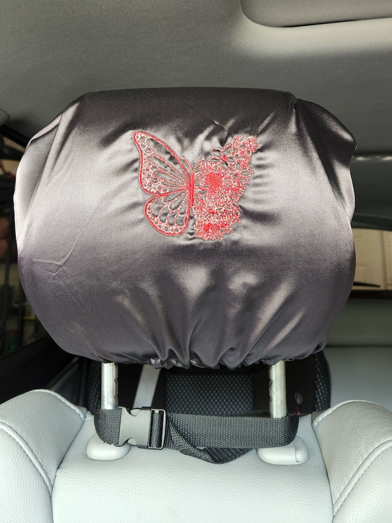 May include: Black satin car headrest cover with a red embroidered butterfly design. The cover has a black strap with a buckle to secure it to the headrest.