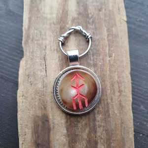 Könnte beinhalten: Silberfarbener Anhänger mit rotem Symbol auf beigem Hintergrund, befestigt an einem silbernen Schlüsselring. Der runde Anhänger liegt auf einem Stück verwittertem Holz. Der Durchmesser des Anhängers beträgt ca. 2,5 cm.