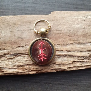 Könnte beinhalten: Ein runder Anhänger mit einem bronzefarbenen Rahmen und einem roten Symbol auf braunem Hintergrund. Der Anhänger hat eine goldfarbene Schlaufe oben. Der Anhänger wird auf einem Stück verwittertem Holz präsentiert.