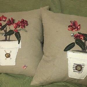 May include: Two beige pillow covers with embroidered floral designs. Each pillow features a white pot with a red flower and green leaves. One pot has a gold button, the other has a striped button.