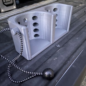 May include: A grey 3D printed double cup holder with a black and white bungee cord attached. The cup holder is sitting in the bed of a truck.