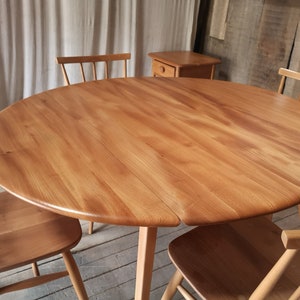 May include: A circular wooden dining table in light brown, with three matching chairs. The table features a split design and a central pedestal. A small wooden side table is in the background. The table's diameter is approximately 120 cm.