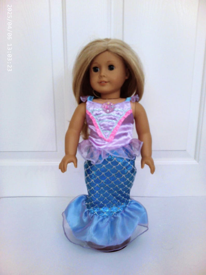 May include: A doll with blonde hair wearing a mermaid costume. The costume features a pink and purple top with a seashell detail and a blue scale-patterned skirt with a ruffled hem. The doll is standing in front of a white background.