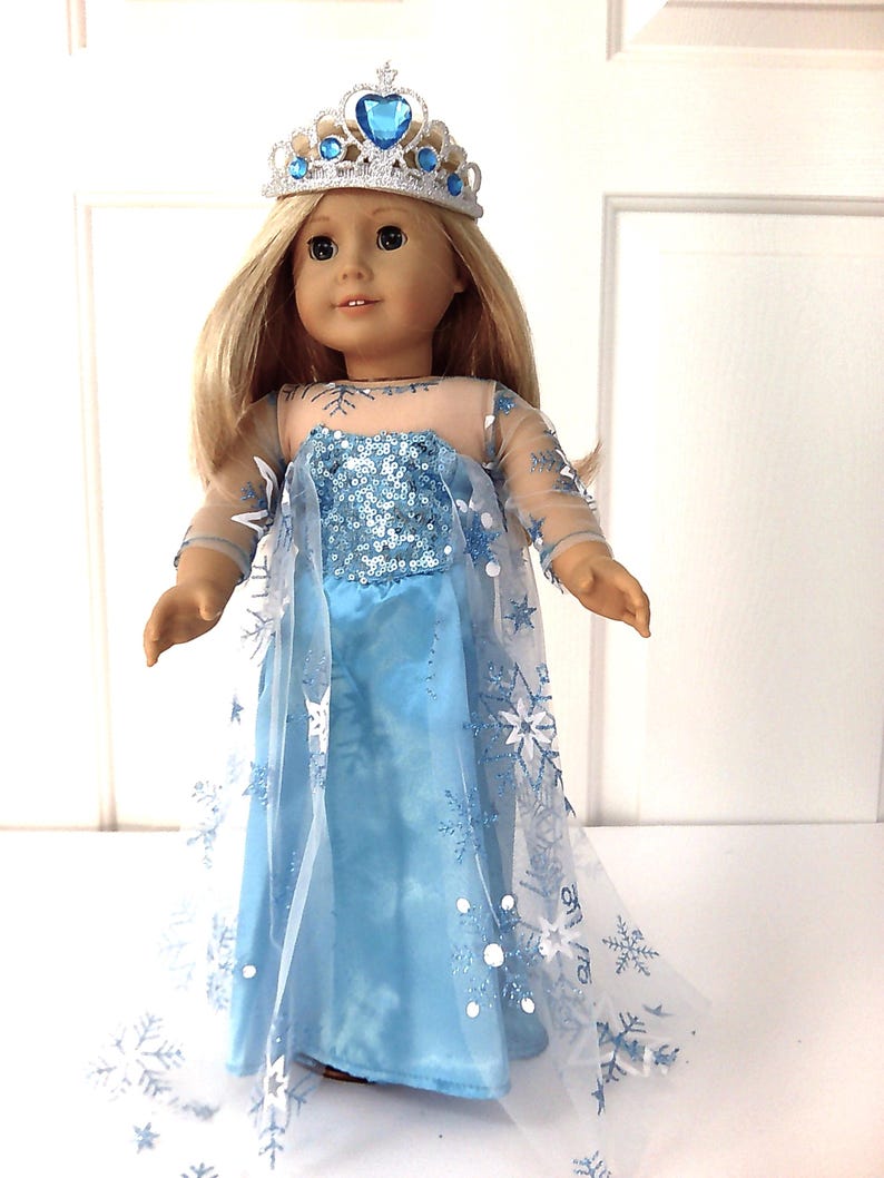 Puede incluir: Mu&ntilde;eca con un vestido azul claro con corpi&ntilde;o de lentejuelas y mangas transparentes. El vestido tiene una falda larga y fluida y una capa transparente adornada con dise&ntilde;os de copos de nieve. La mu&ntilde;eca tambi&eacute;n lleva una tiara plateada con detalles azules.