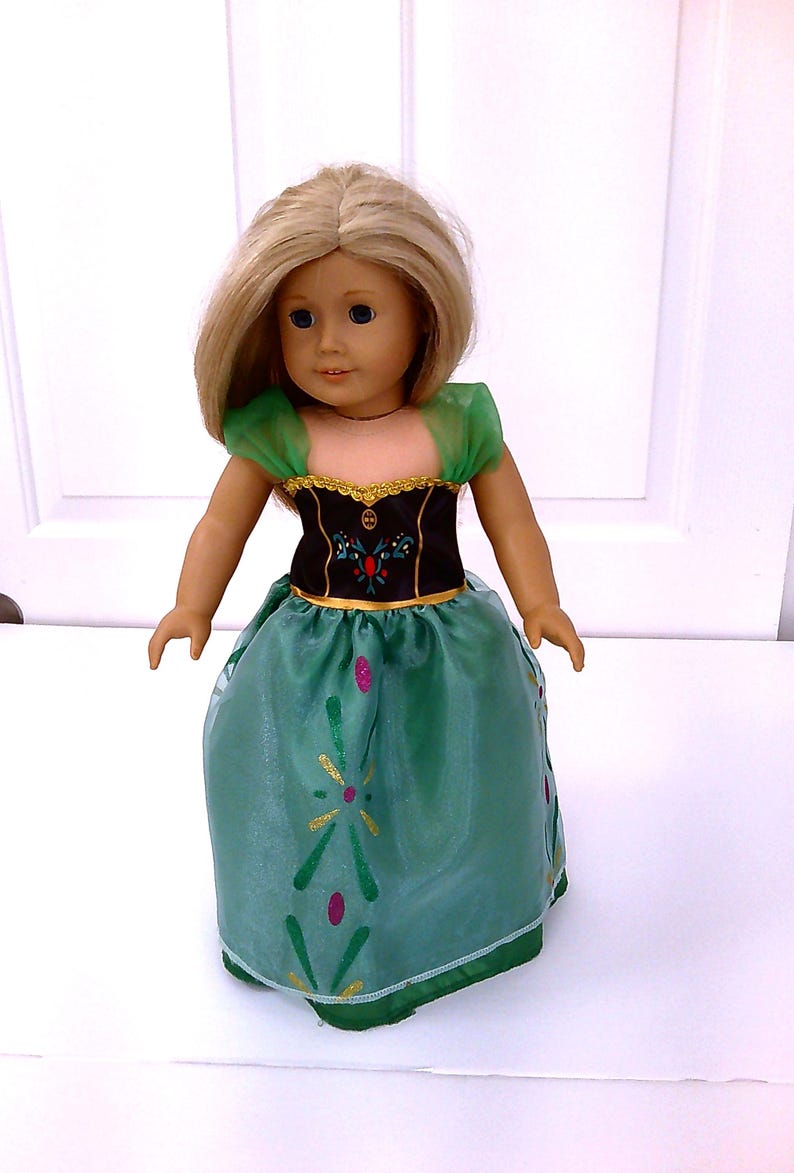 May include: A doll with blonde hair wearing a green and black dress. The dress has a green skirt with gold and pink accents. The bodice is black with gold trim and a floral design. The doll has blue eyes and is standing.