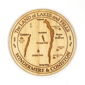 May include: A wooden coaster with a map of the Lake District in England. The map shows the locations of Coniston, Windermere, Ambleside, and other points of interest. The coaster is engraved with the text "The Land of Lakes and Fells, Windermere & Coniston".