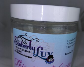 Op de afbeelding: Een doorzichtige plastic pot met een witte deksel, met het label "Sisterly Luv Moisturizing Cream" voor alle haartypes. Het label luidt "Thirsty for Moisture" met een stripfiguur. De pot bevat een romige substantie.