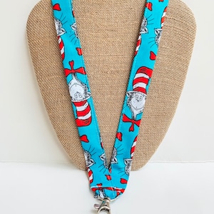 May include: A blue lanyard featuring a repeating pattern of a cartoon cat wearing a red and white striped hat and bow tie. The lanyard has a silver-colored clasp and key ring.