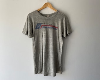 May include: Gray short-sleeved t-shirt with a red, white, and blue graphic that says "Fit to Win".