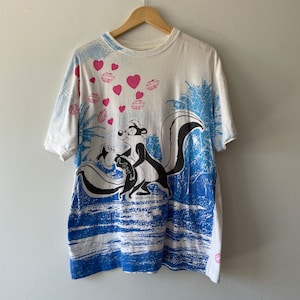 Puede incluir: Camiseta blanca con una imagen de dibujos animados de Pepe Le Pew y Penelope Pussycat. La camiseta tiene un patrón de olas azul y blanco y corazones y labios rosas.