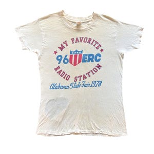May include: A white short-sleeve t-shirt featuring a graphic with the text MY FAVORITE 96 MERC RADIO STATION and Alabama State Fair 1978. The design includes a red and blue shield emblem with stars. There is a dark grey stain on the lower left side.