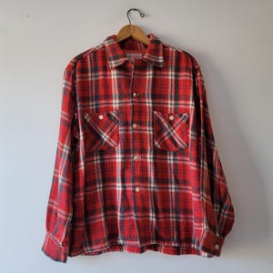 Pode incluir: Camisa de flanela xadrez vermelha e preta de manga comprida e fecho de botões. A camisa tem dois bolsos no peito com fecho de botões.
